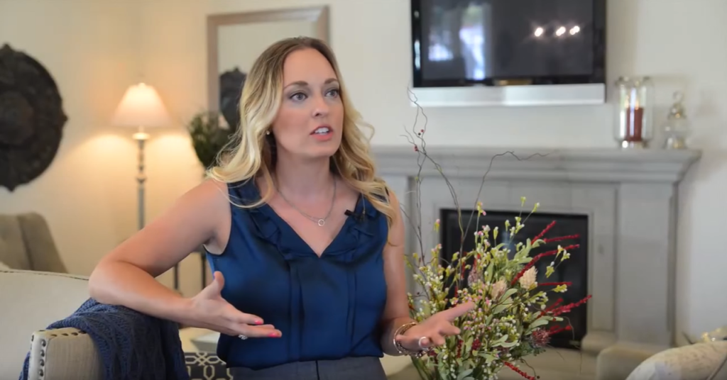 A woman in a blue top gestures while speaking, sitting in a well-furnished room with a fireplace and a muted television in the background.