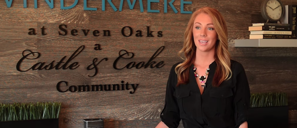 A woman with long red hair, wearing a black blouse and a necklace, stands smiling in front of a wooden wall with the sign "windermere at seven oaks, a castle & cooke community.