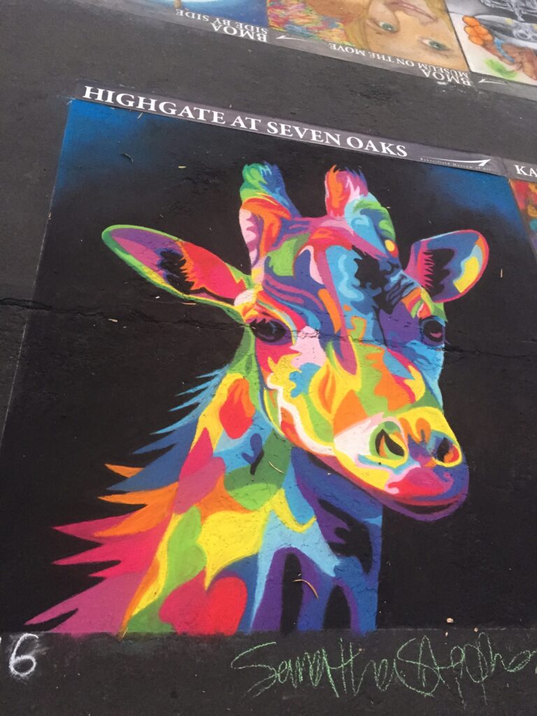 Colorful street art depicting a zebra painted in vibrant, multi-colored hues on a dark urban wall, signed by the artist at the bottom.