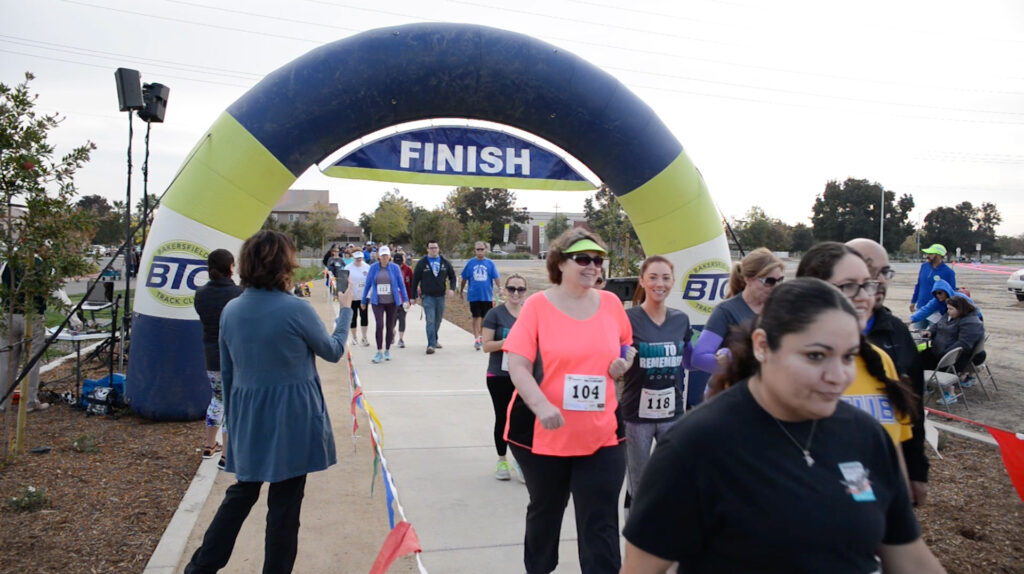 Participants crossing the finish line under a large blue and yellow inflatable arch at a community race event, with spectators on the sides.