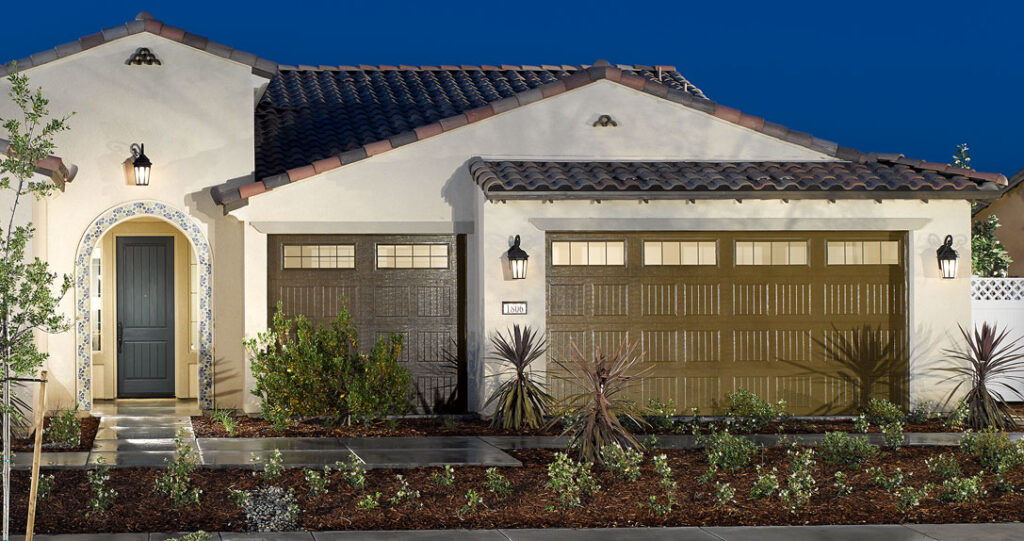 Modern single-story house with a tiled roof and a beige stucco exterior, featuring a garage and a landscaped front yard with small trees and desert plants.