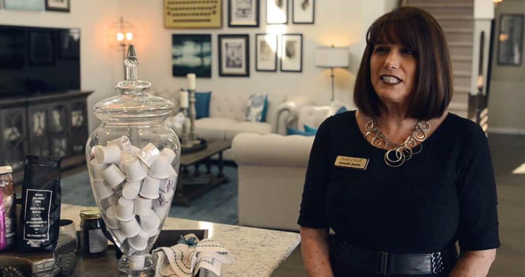 A woman with brown hair, wearing a black outfit and a name tag, stands in a stylish room with framed pictures, a large jar filled with scrolls, and elegant furniture.