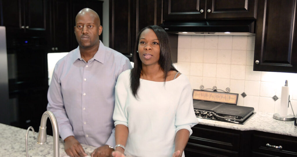 A man and a woman standing in a modern kitchen with white cabinets and black countertops. the woman is speaking and gesturing while the man stands next to her looking at the camera.