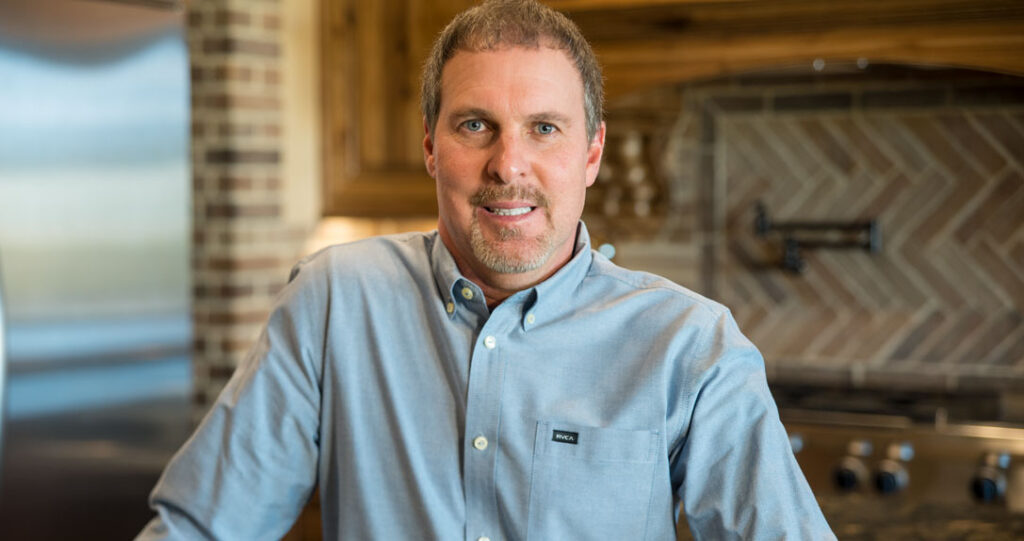 A man with light skin and short gray hair wearing a blue shirt smiles while standing in a kitchen with wood cabinets and a stylish backsplash.