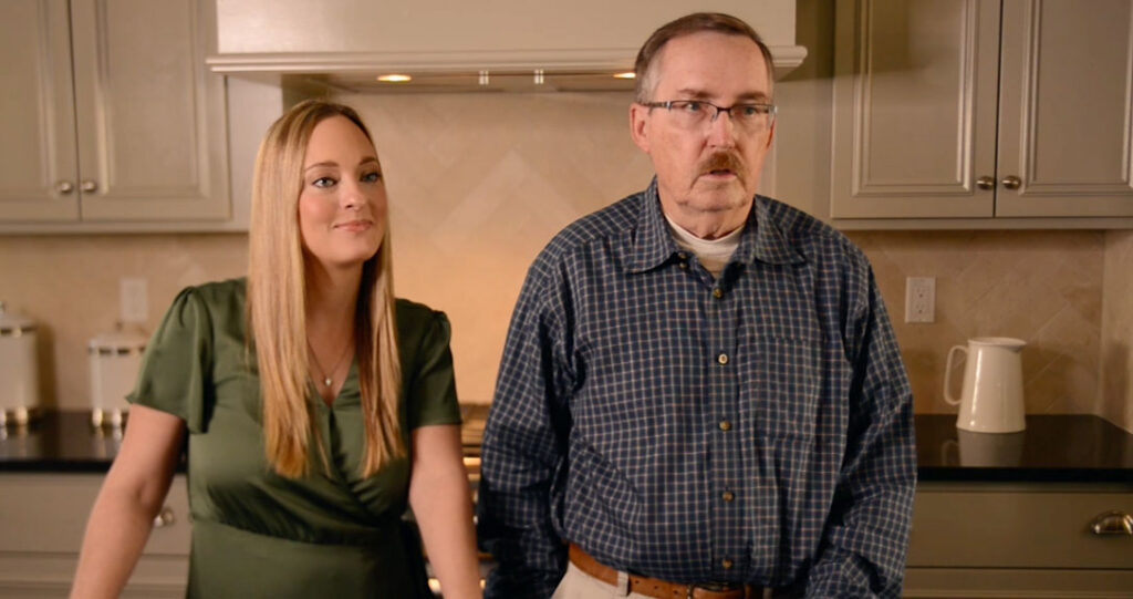 A man and a woman standing in a kitchen with modern decor. the woman appears to be smiling subtly, while the man looks straightforward with a neutral expression.