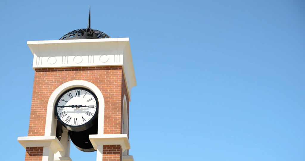 A traditional clock tower with a white face and black roman numerals stands against a clear blue sky, framed by a brick structure with a pointed arch.