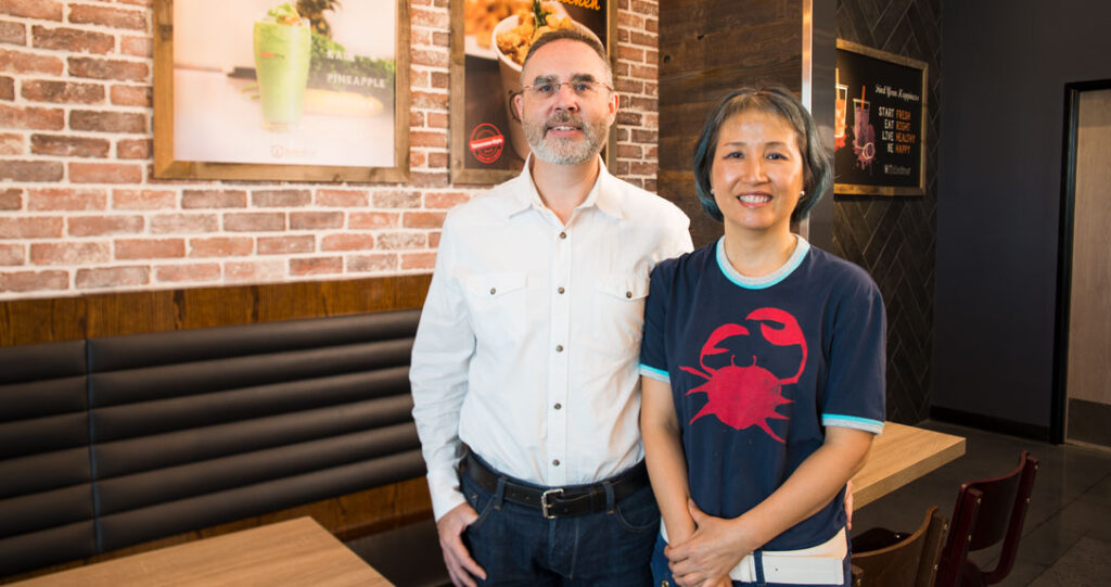 A man and a woman smiling inside a restaurant, with the man wearing glasses and a white shirt, and the woman in a blue t-shirt with a red crab design, standing in front of a brick wall and a black booth.