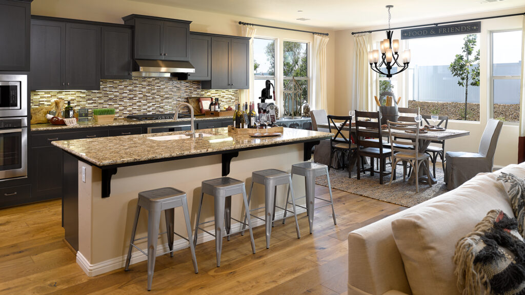 Spacious modern kitchen with dark cabinetry, stainless steel appliances, and a large island, extending to a dining area with a wooden table set and elegant chandelier.