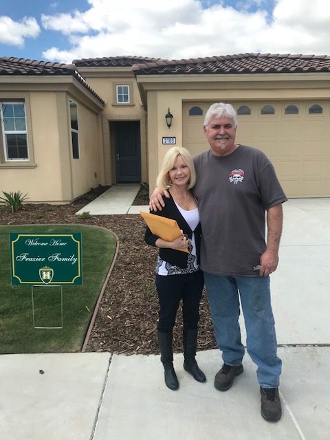 A mature couple smiling in front of their new tan stucco home with a green front lawn and a sign that reads "welcome home frontier family.