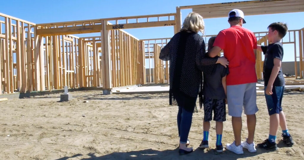 A family of four, viewed from behind, stands at a construction site observing the wooden framework of a building on a sunny day.