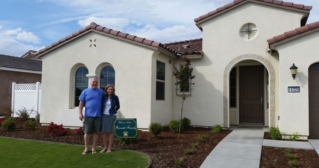 A smiling older man and a younger woman standing in front of a new cream-colored house with a spanish tile roof, holding a sign that says "sold." there's a lush green lawn and a clear blue sky.