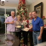 Five adults standing and smiling in a living room decorated for christmas, holding dessert plates, with a tall, elaborately decorated tree in the background.