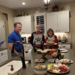 Three adults stand in a well-lit kitchen with a spread of food on the island, smiling and enjoying a casual gathering. decor suggests a cozy, festive atmosphere.