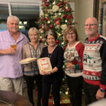 Five adults smile beside a large, ornately decorated christmas tree, holding drinks and festive treats. two wear christmas sweaters. warm indoor setting.