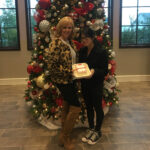 Two women smiling, standing in front of a large, decorated christmas tree, holding a festive cake. one woman is in a leopard print jacket and boots, the other in a black coat and sneakers.