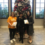 Three women smiling and posing with holiday cakes in front of a large, ornately decorated christmas tree inside a building.