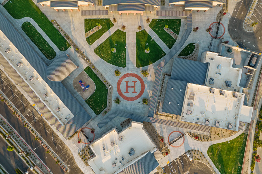 Aerial view of a hospital complex with helipad marked with an "h", landscaped pathways, surrounding roads, and parking spaces, captured during daylight.