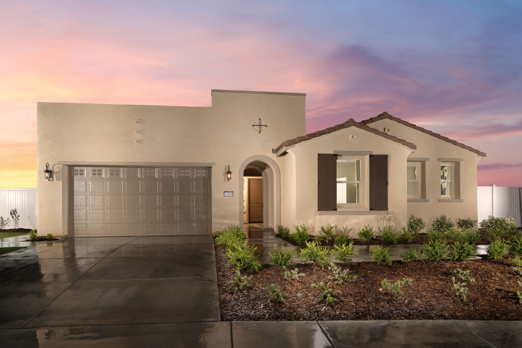 A single-story suburban home with a beige stucco finish and a two-car garage at dusk. the house features lush landscaping and a dramatic colorful sky in the background.