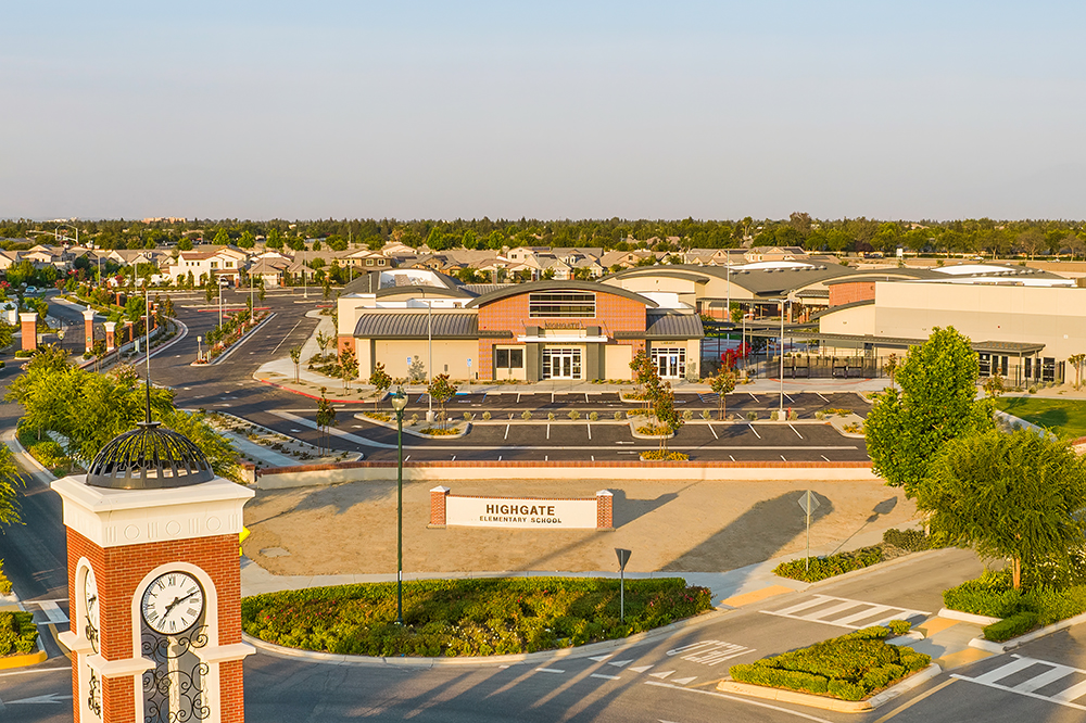 Aerial view of a suburban area featuring a clock tower with the inscription "highgate" at the foreground, surrounding roads, parking lots, and residential housing under a clear sky.