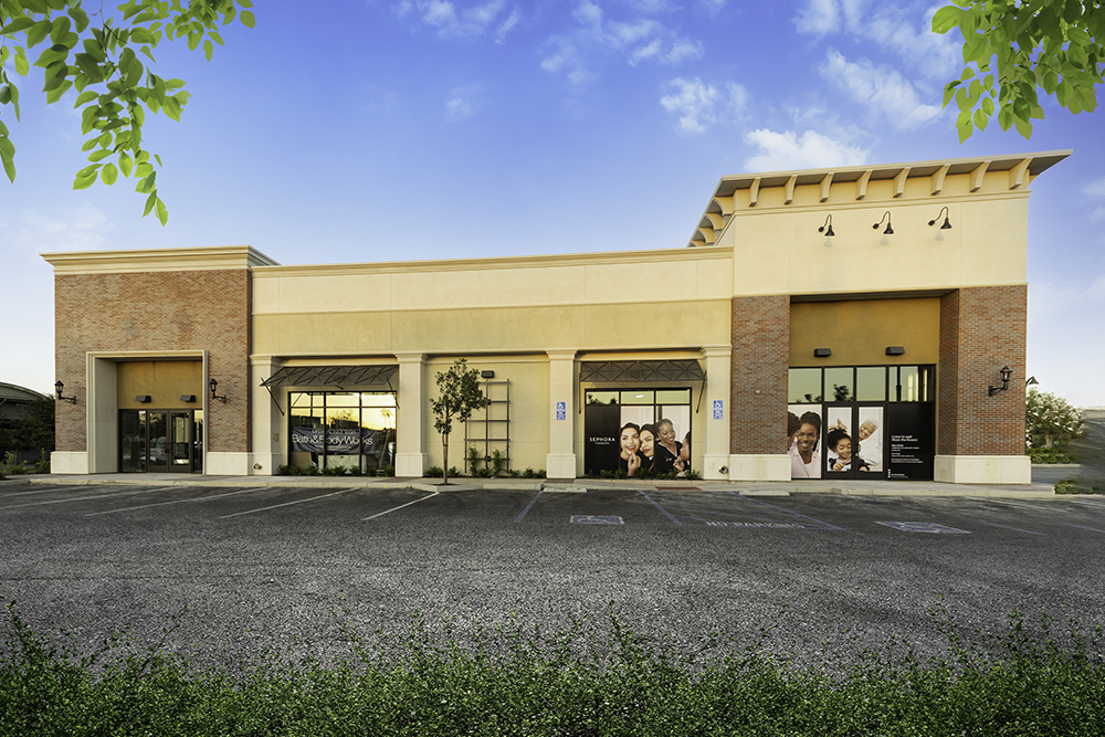 A modern single-story commercial building with a tan facade, large glass windows displaying posters, and a spacious parking lot in front, set against a clear blue sky.