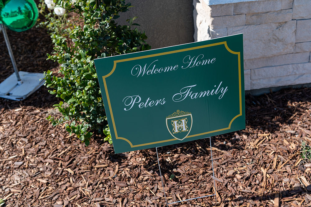 A welcome sign for the "peters family" posted in a garden with a green background, decorative white border, and a family crest, surrounded by brown mulch.