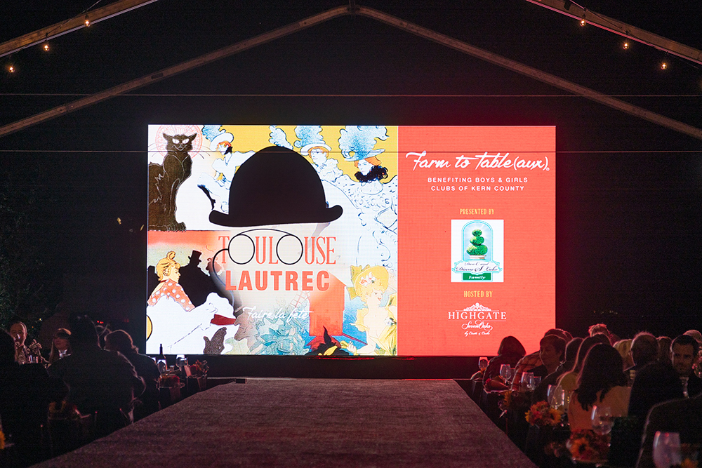 A large digital screen displays a toulouse-lautrec themed graphic at a charity event, with guests seated under a tent and a banner promoting a benefit for boys & girls clubs.