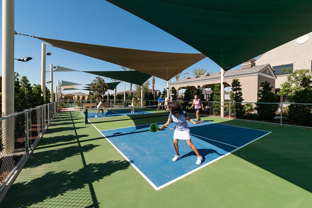 People playing pickleball on outdoor courts shaded by canopies on a sunny day, with residential buildings in the background.