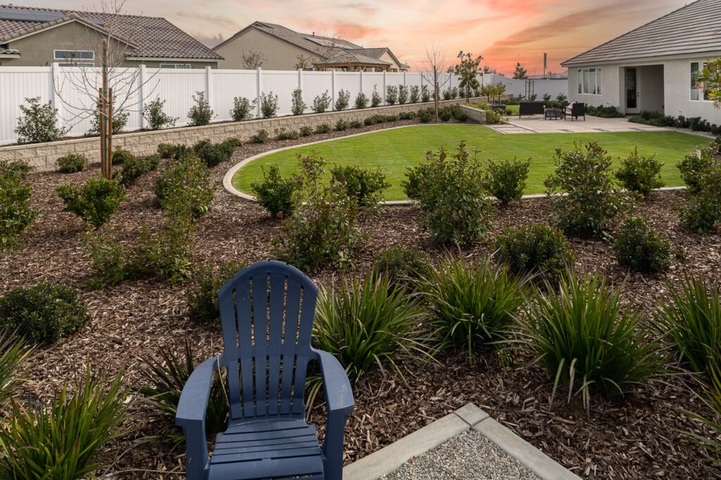 A serene backyard garden at sunset with a winding path, manicured lawn, and surrounded by lush shrubbery and plants. a blue chair stands in the foreground, facing a distant patio area.