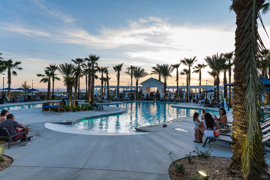 A tranquil poolside scene at dusk with tall palm trees, reclined visitors, and a clear blue pool reflecting the evening sky, set in a resort-like setting.