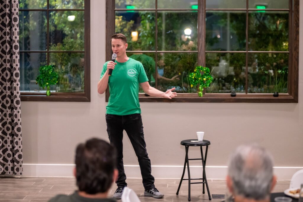 A man in a green t-shirt stands holding a microphone, addressing an audience in a room with large windows and decorative green lights.