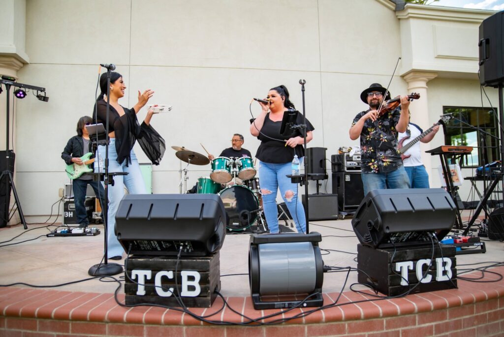 A lively band performs outdoors on a small stage. two female vocalists joyfully sing into microphones while accompanied by a guitarist, a drummer, and a violinist.