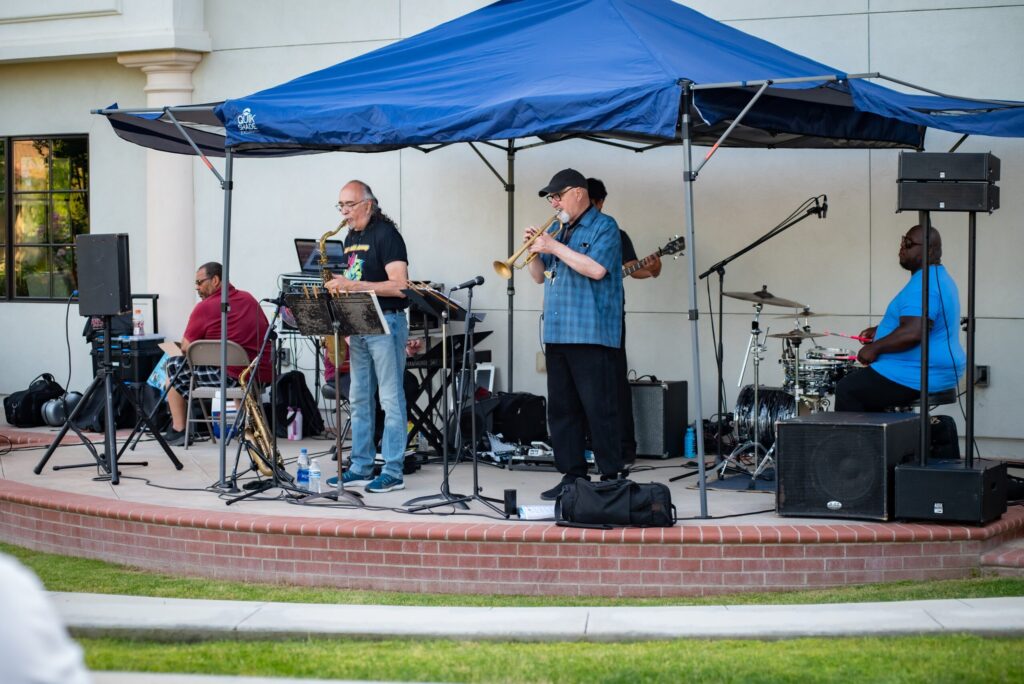 A band consisting of four members performs outdoors under a blue canopy. they include a keyboardist, flutist, guitarist, and a drummer, playing on a sunny day near a building.