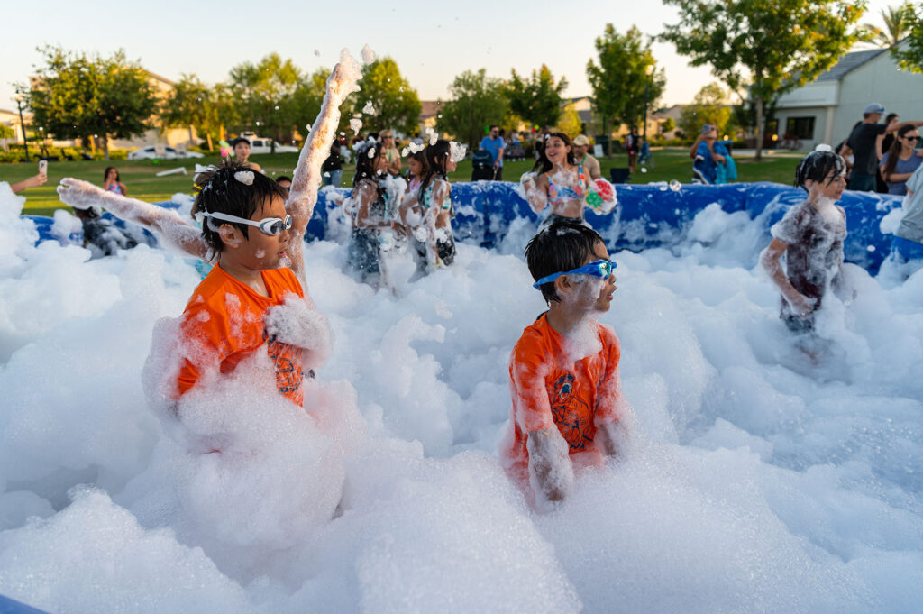 Children wearing goggles and orange shirts, joyfully playing in a large foam-filled pool at an outdoor event, with trees and a clear sky in the background.