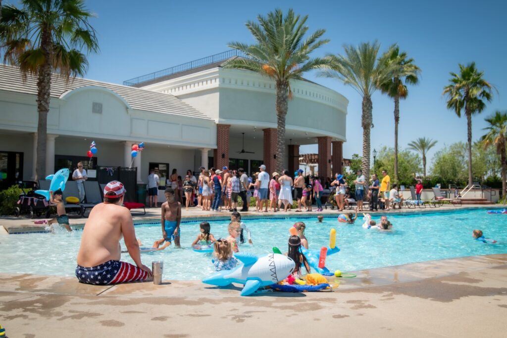 A vibrant outdoor pool scene at a community center on a sunny day, featuring people of various ages swimming and socializing, with palm trees and a clear blue sky in the background.