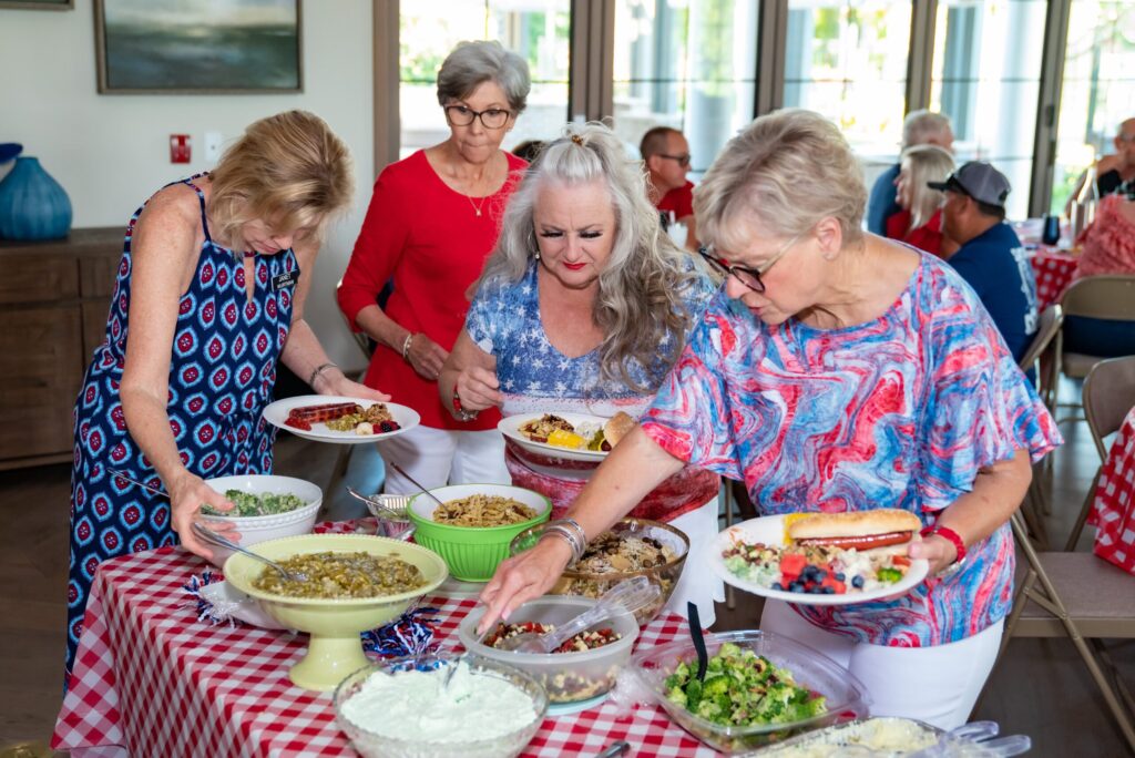 Four older women serving themselves at a potluck with various dishes on a table covered in a red-checked tablecloth, inside a bright, cozy room.