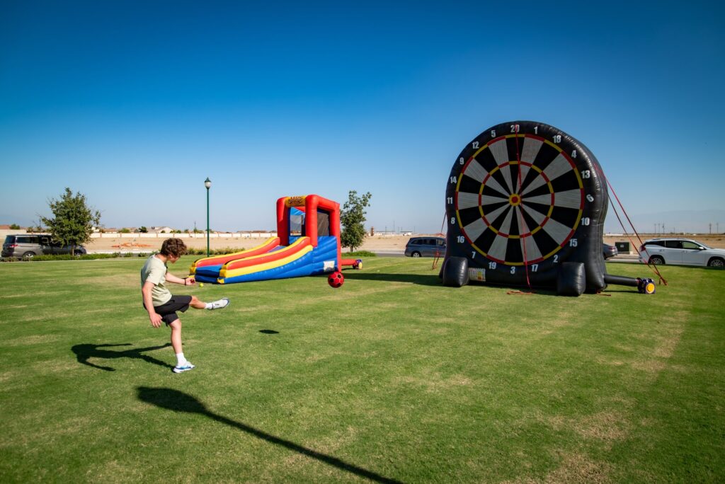 A person kicks a soccer ball towards a giant inflatable dartboard in a grassy park with a colorful bounce house and parked cars in the background.