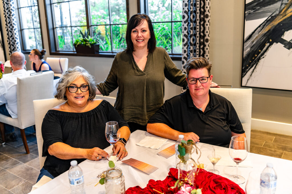 Three women smiling at a dining table in a refined restaurant setting, with one standing behind the other two who are seated. glasses of wine and elegant decor visible.