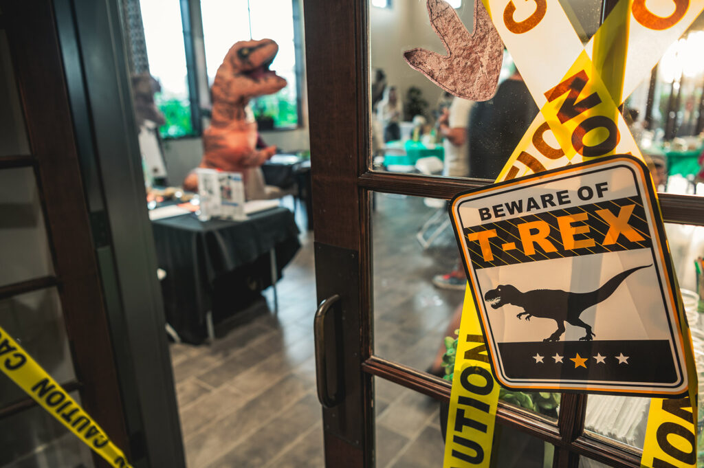 A signage saying "beware of t.rex" is displayed in the foreground with a blurry image of a person in a dinosaur costume visible through a glass door at a themed event.