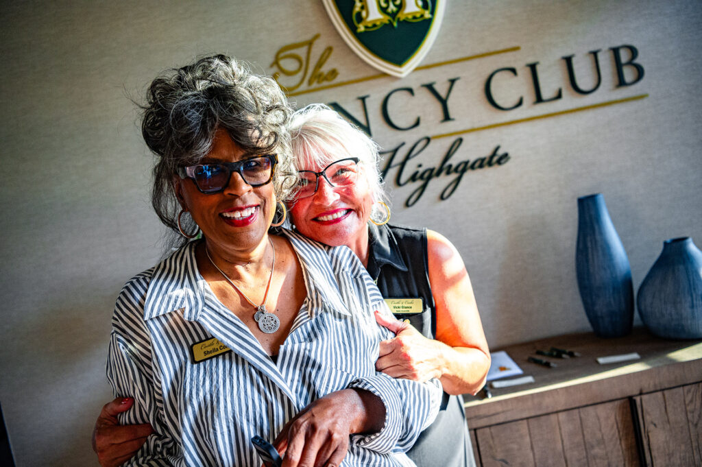 Two smiling women wearing name tags embracing in front of a sign that reads "the legacy club highgate." they appear joyful and are dressed in professional attire.