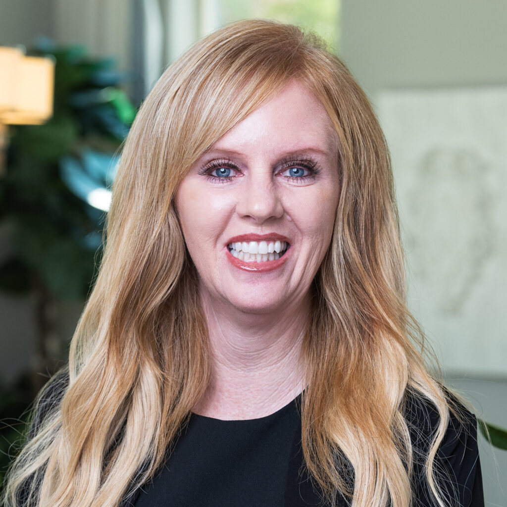 Portrait of a smiling woman with long blonde hair and fair skin, wearing a black blouse, standing in an office setting with green plants and a map in the background.