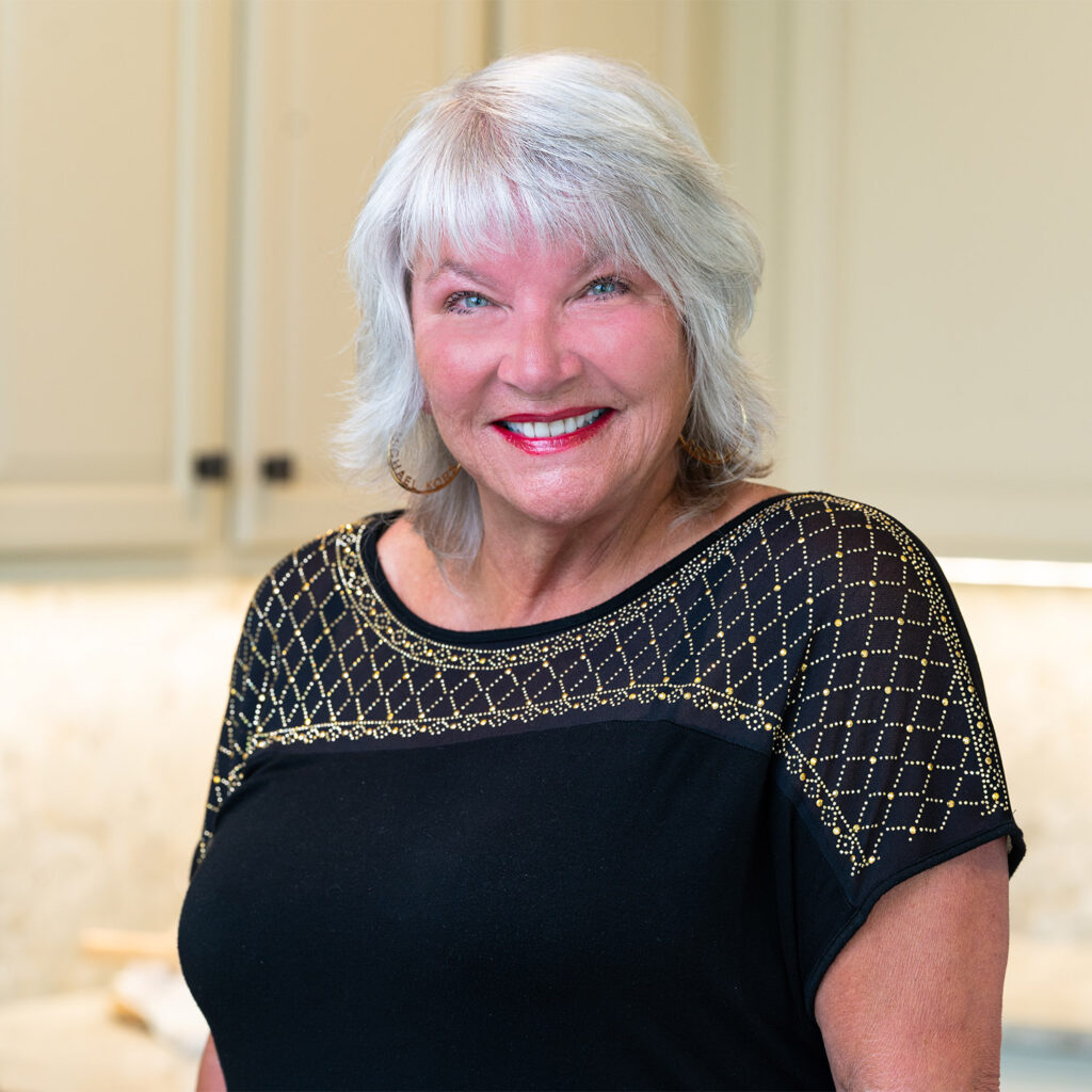 A smiling elderly woman with short white hair, wearing a black top with gold embellishments, posed in a kitchen setting.