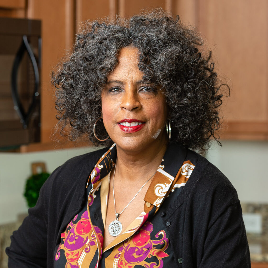 A smiling woman with curly gray hair, wearing a colorful blouse and a necklace, stands in a kitchen with wooden cabinets in the background.