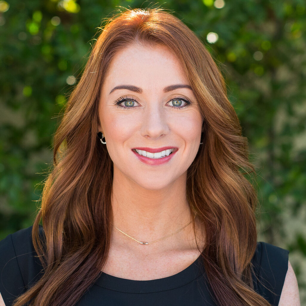 A professional headshot of a woman with long red hair, smiling, wearing a black top and subtle gold necklace, standing against a blurred greenery background.