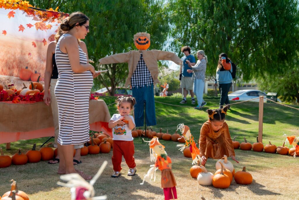 A lively outdoor pumpkin patch scene with a child in a pumpkin costume, a woman, and a toddler exploring fall decorations, surrounded by other visitors on a sunny day.