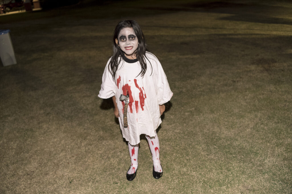 A young child dressed in a spooky halloween costume, featuring a white shirt with red blood-like stains, dark eye makeup, and black lipstick, holding a toy weapon, standing on grass at night.