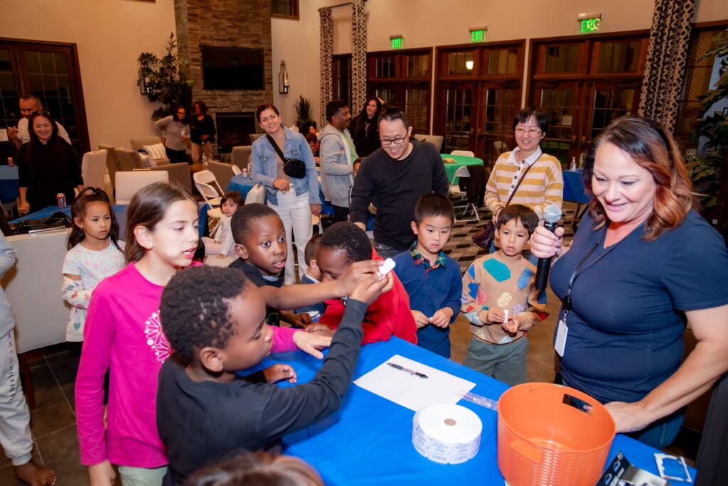 A diverse group of excited children surround a table at a community event, engaging in a science experiment led by a smiling woman, with other adults observing in the background.