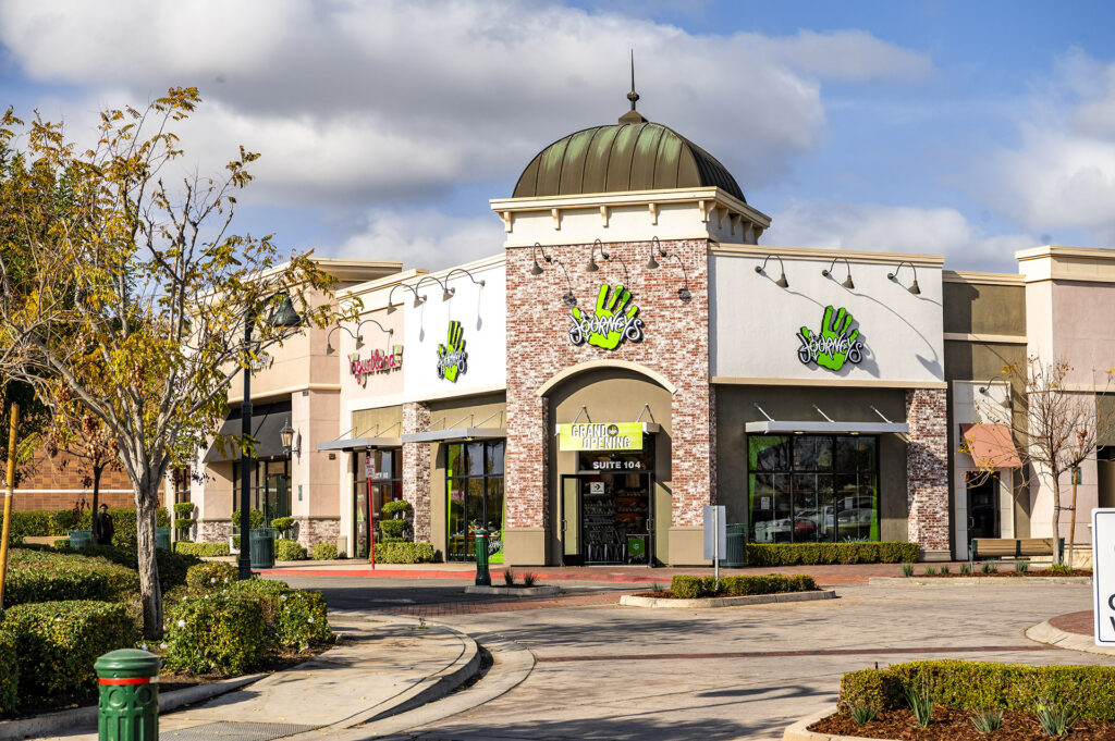 View of a shopping center with a store named "hand & stone" featuring vibrant green handprints on beige walls. the building has a domed structure on the roof, trees, a sunny sky, and a paved driveway.