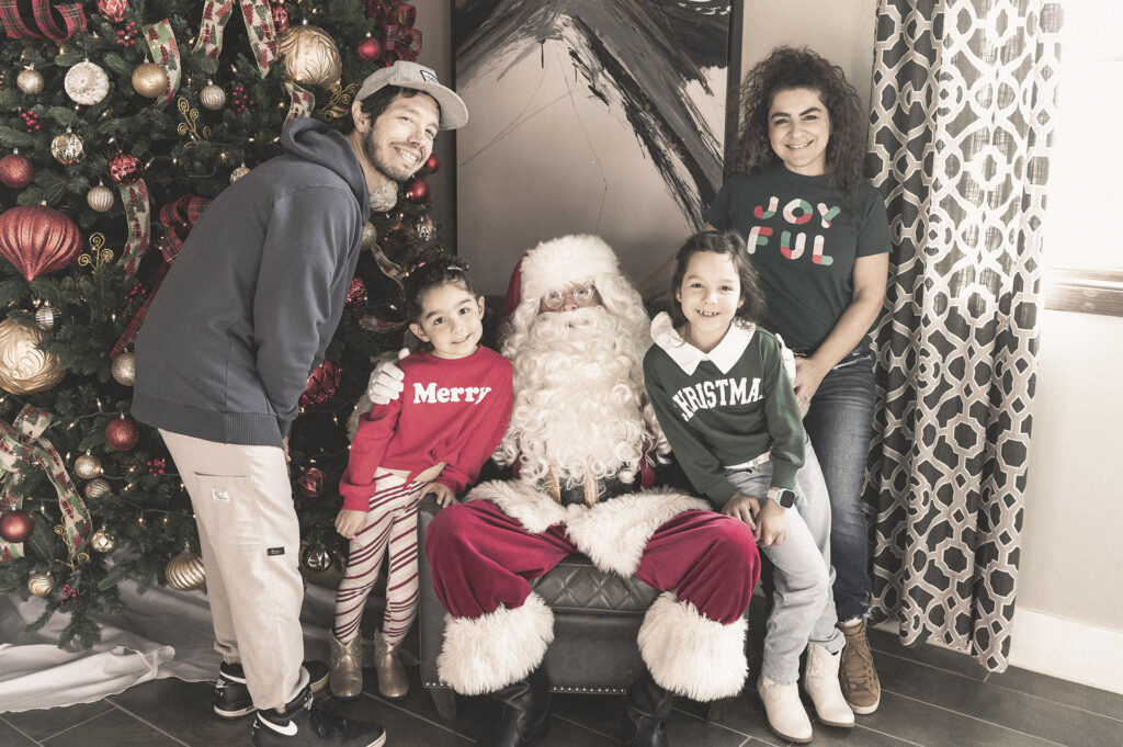 A family of four, two adults and two children, posing happily with santa claus by a christmas tree decorated with red and gold ornaments.