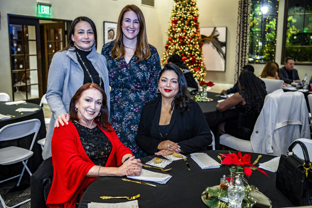 Four women smiling at a holiday party, with two standing and two seated at a table adorned with festive decorations and a christmas tree in the background.