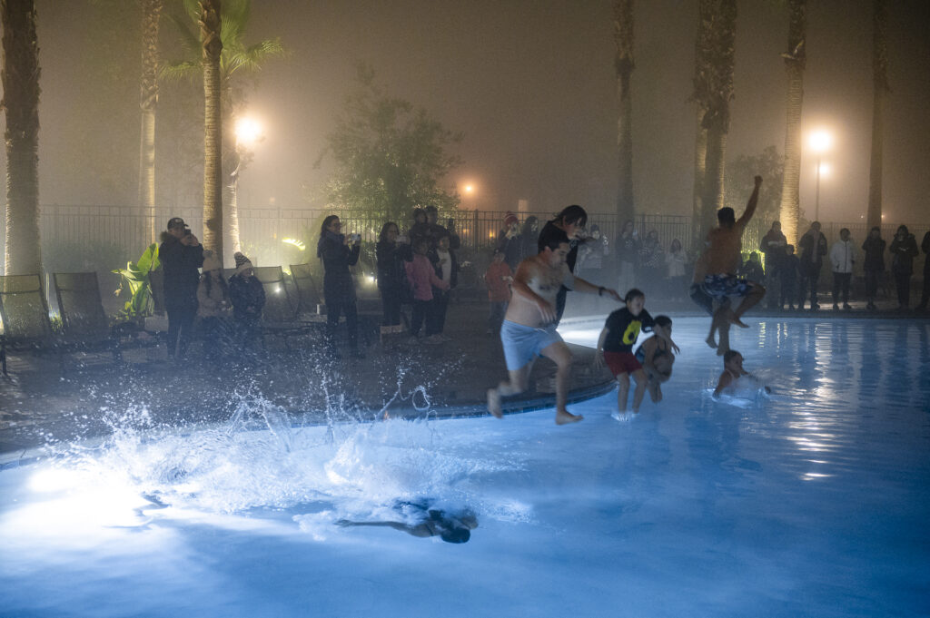 People enjoy a nighttime pool party amidst heavy fog. some individuals perform playful jumps into the illuminated water, creating splashes, while onlookers watch from the poolside, wrapped in towels or casual wear.